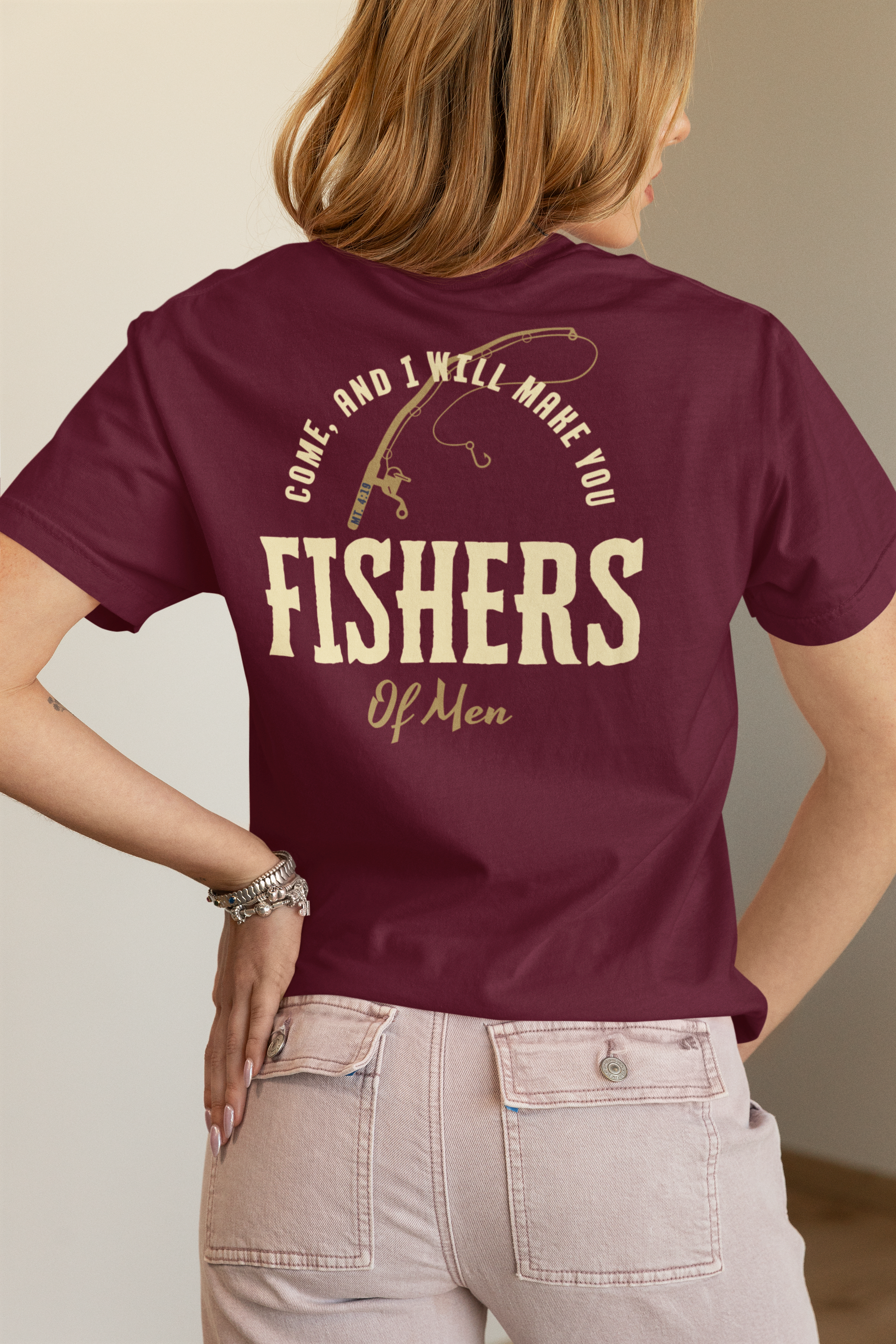 A woman shown from the back wearing a maroon Christian graphic T-shirt with a beige fishing rod illustration and the words “COME, AND I WILL MAKE YOU FISHERS OF MEN,” paired with light beige jeans.