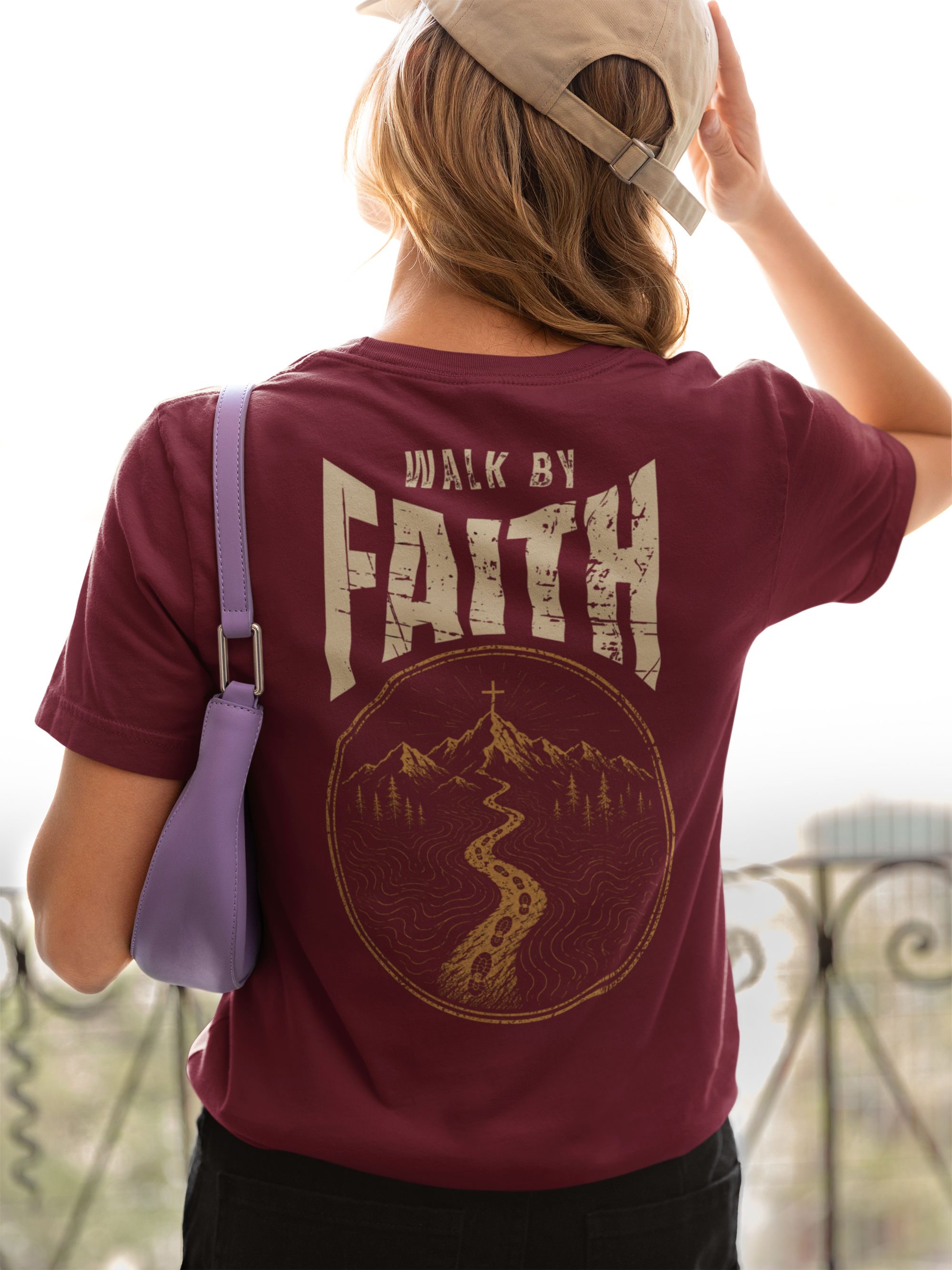 A woman viewed from behind wearing a maroon Christian outdoor t-shirt with a large distressed graphic on the back that reads 'WALK BY FAITH' above a mountain and path illustration. She is wearing a beige cap and carrying a purple shoulder bag.