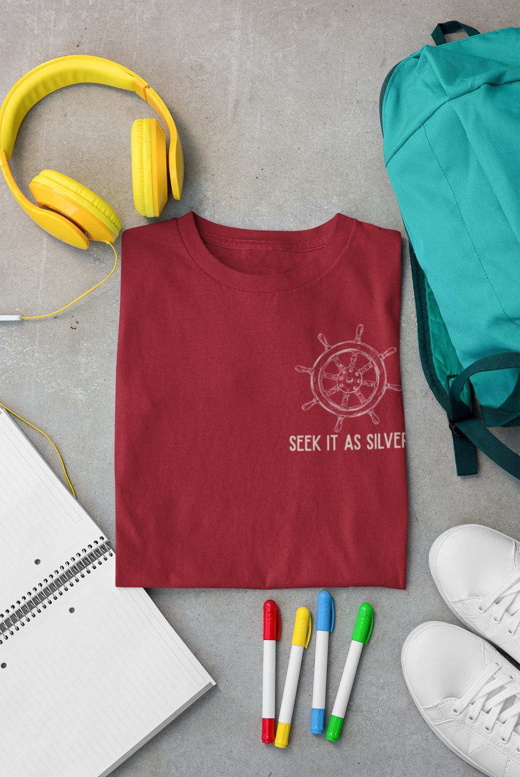 A folded maroon bible verse t-shirt with a white ship's wheel graphic and the text 'SEEK IT AS SILVER' is displayed in a flat-lay arrangement on a gray concrete background, surrounded by yellow headphones, a teal backpack, a notebook, colored markers, and white sneakers.