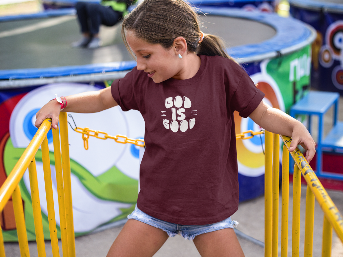 A young girl with her hair in a ponytail smiles while leaning on yellow playground railings at an outdoor fair. She is wearing a maroon Christian graphic t-shirt by SeedSowerGraphics with a white graphic that reads 'GOD IS GOOD' and denim shorts.