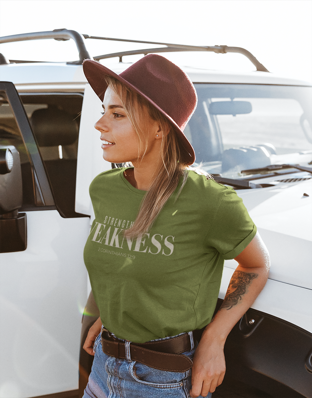 A woman wearing a military green Christian graphic t-shirt and a brown felt hat leans against a white vehicle outdoors. The t-shirt features the text 'STRENGTH IN WEAKNESS' in white lettering.