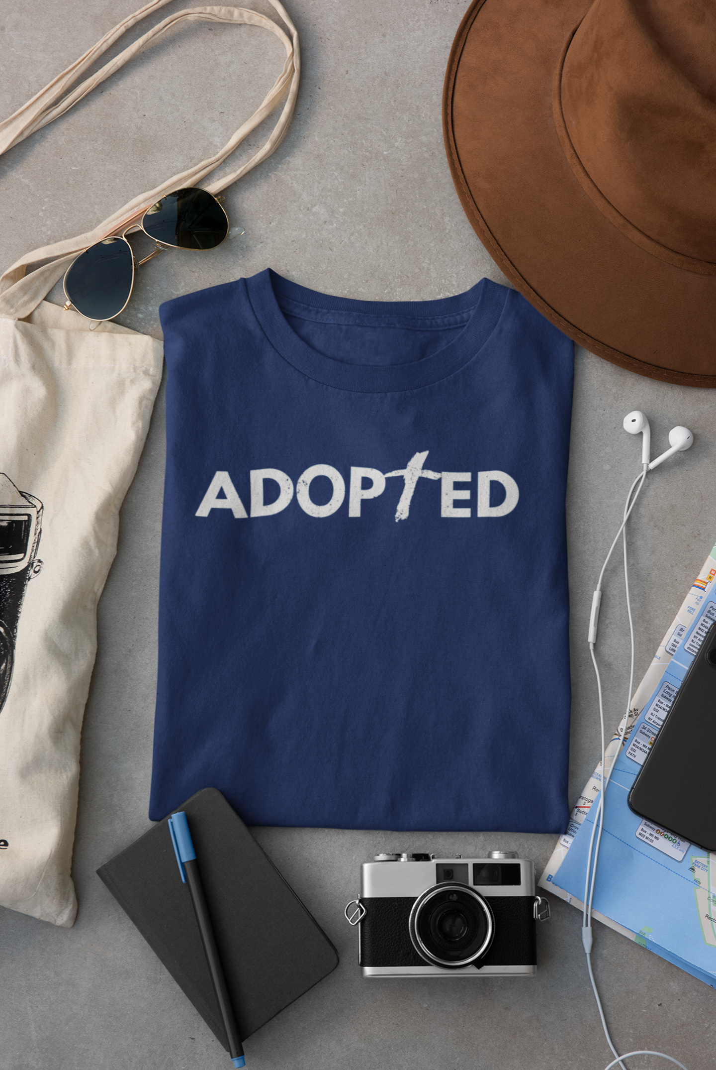 Navy Blue folded Christian T-shirt that says 'Adopted' surrounded by travel accoutrements such as sunglasses, camera, headphones, map, and a brown hat.