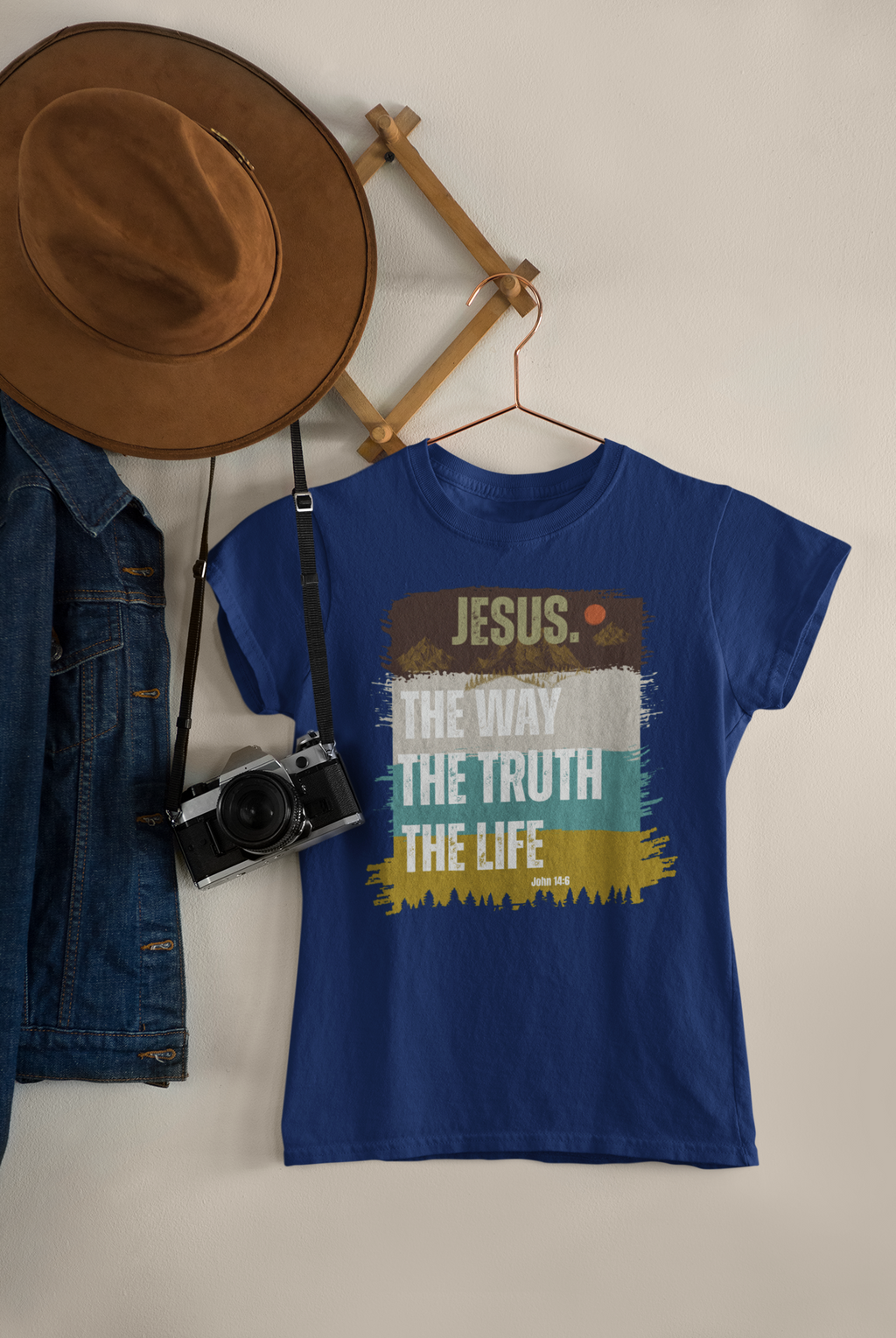 A royal blue Christian graphic t-shirt hanging on a wooden hanger against a light beige wall. The shirt features a distressed graphic with the word 'JESUS. The Way the truth the life."