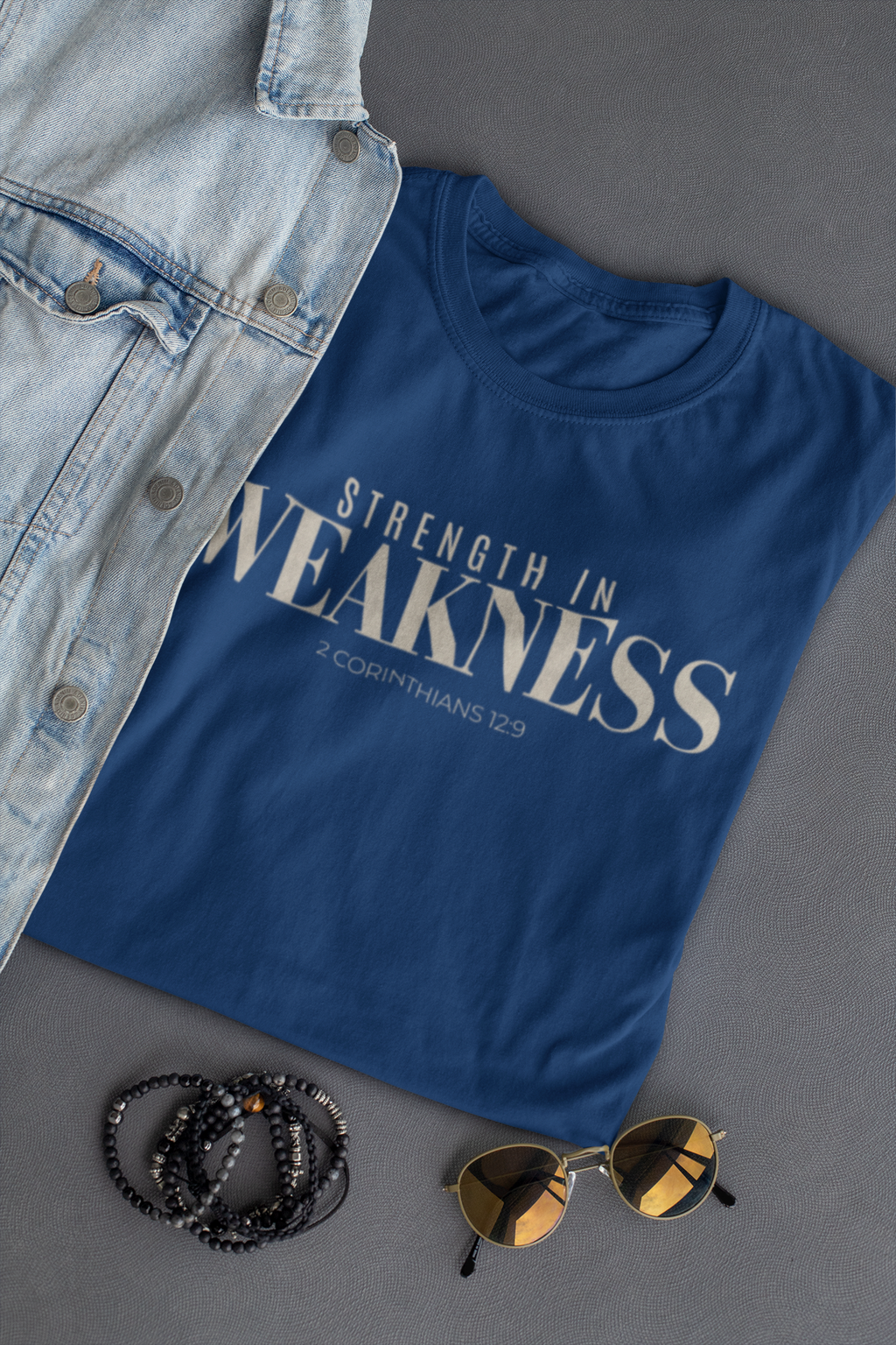 A folded navy Christian graphic T-shirt with the phrase 'STRENGTH IN WEAKNESS 2 CORINTHIANS 12:9' in white text, styled in a flat lay with a light denim jacket, black beaded bracelets, and round sunglasses on a dark gray background.