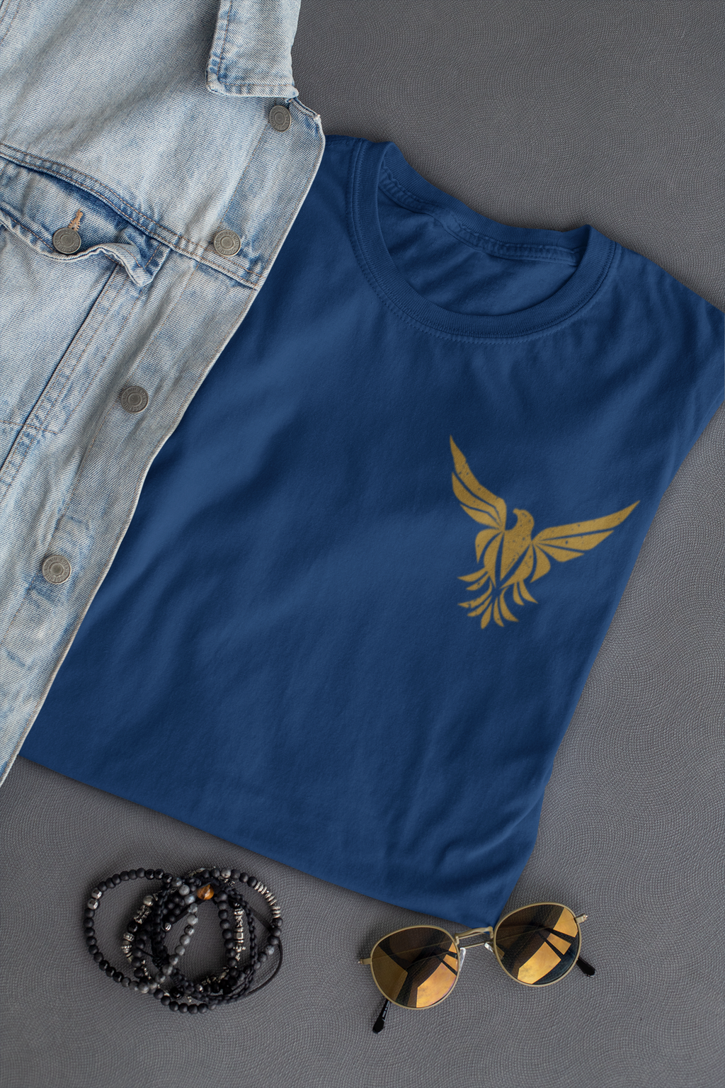 A navy blue Christian graphic t-shirt with a gold phoenix graphic on the left chest, styled in a flat-lay with a light-wash denim shirt, black beaded bracelets, and sunglasses on a dark gray background.