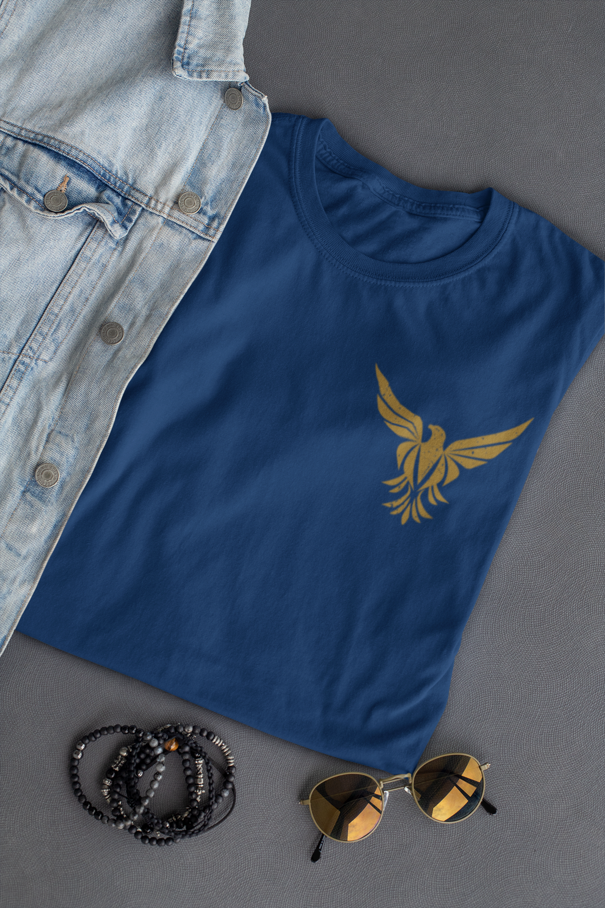 A navy blue Christian graphic t-shirt with a gold phoenix graphic on the left chest, styled in a flat-lay with a light-wash denim shirt, black beaded bracelets, and sunglasses on a dark gray background.