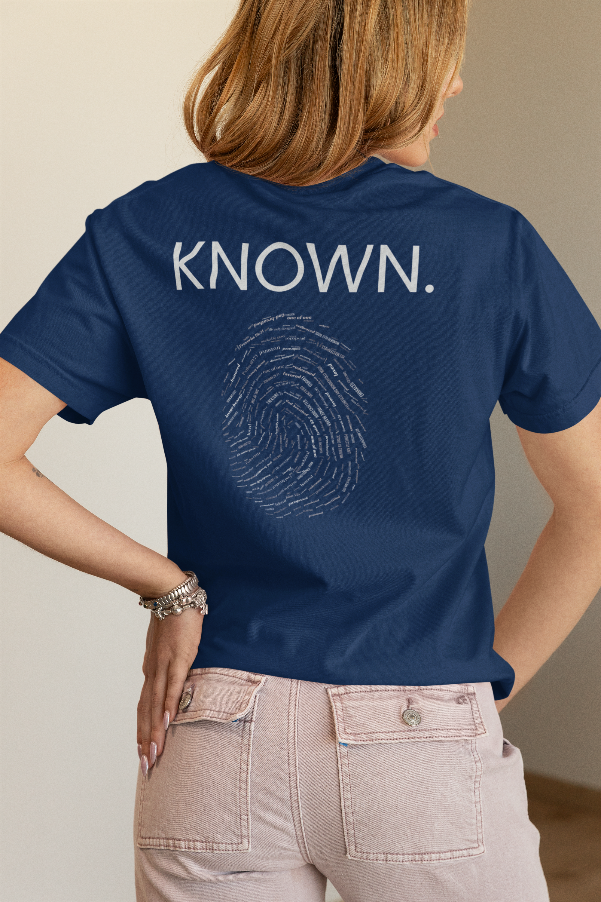 A woman shown from the back wearing a navy blue Christian Bible verse t-shirt with 'KNOWN.' and a fingerprint graphic printed in white on the back, paired with light beige jeans.
