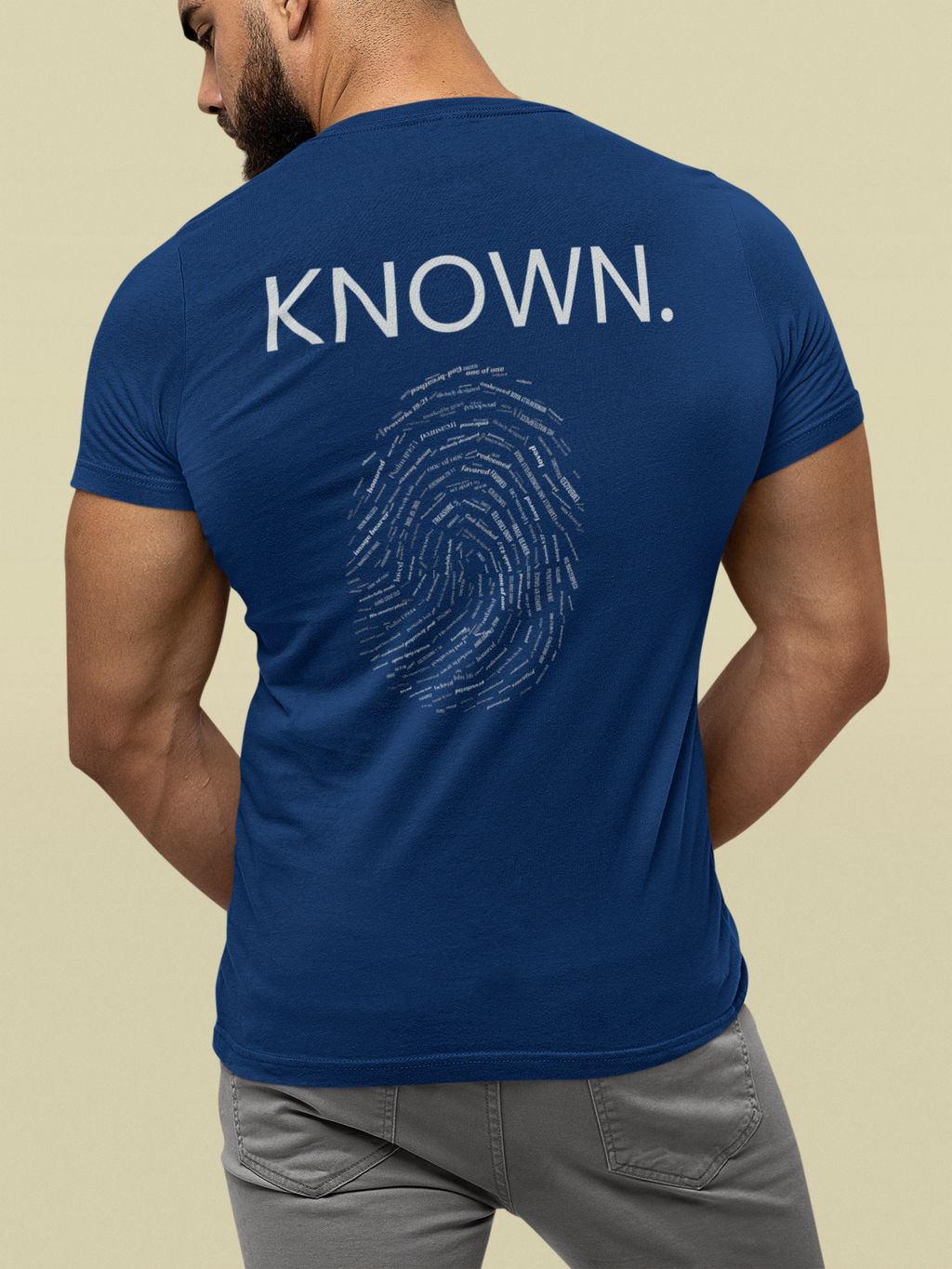 A man viewed from the back wearing a royal blue Christian graphic t-shirt with 'KNOWN.' printed in white above a white fingerprint graphic with words of affirmation inlaid, paired with gray pants against a solid beige background.