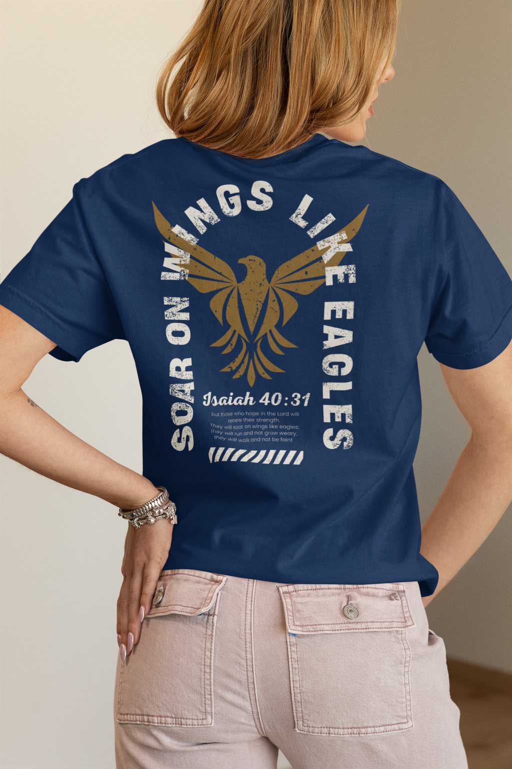A woman shown from the back wearing a navy blue Christian graphic t-shirt with a large gold eagle graphic and white text reading 'Soar ON WINGS LIKE EAGLES' and 'Isaiah 40:31'. She is wearing light pink jeans against a neutral beige background.