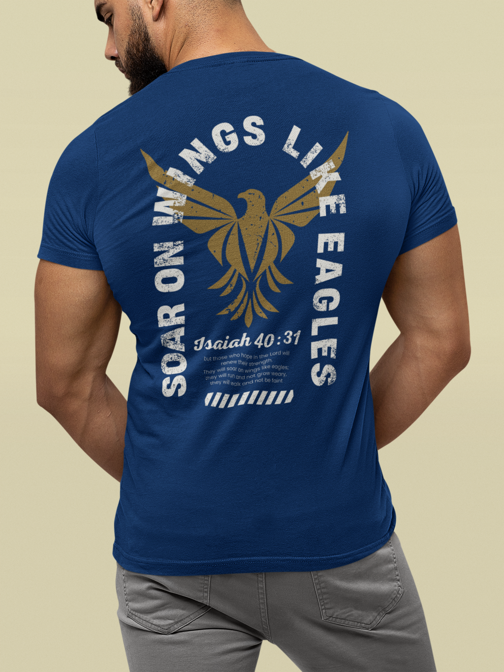 A man wearing a navy Bible verse t-shirt from the back, featuring a large graphic print of an eagle with wings like eagles, the text 'SOAR ON WINGS LIKE EAGLES', and 'Isaiah 40:31' in white and gold. He is wearing gray pants against a solid beige background.