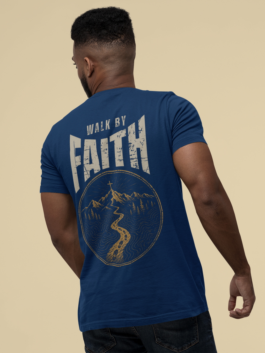 A man shown from the back wearing a royal blue Christian graphic t-shirt with a distressed graphic on the back that reads 'WALK BY FAITH' above a circular mountain and road illustration. The photo is set against a solid beige background.
