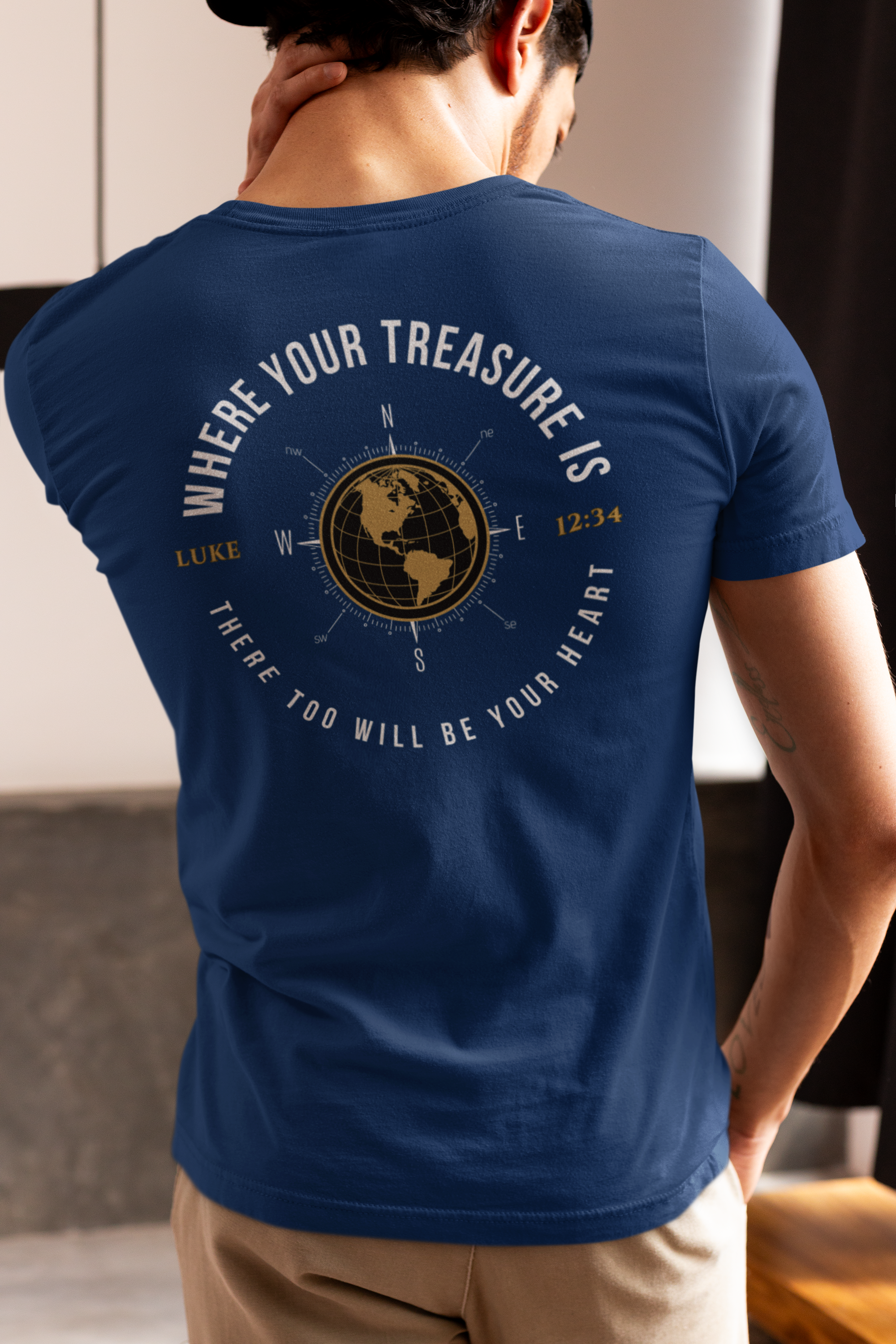 A man viewed from behind wearing a navy blue Christian Bible verse t-shirt with a large circular graphic on the back. The graphic features a gold globe compass, the text 'WHERE YOUR TREASURE IS' arched above, and 'THERE TOO WILL BE YOUR HEART' curved below, along with 'LUKE 2:34'.