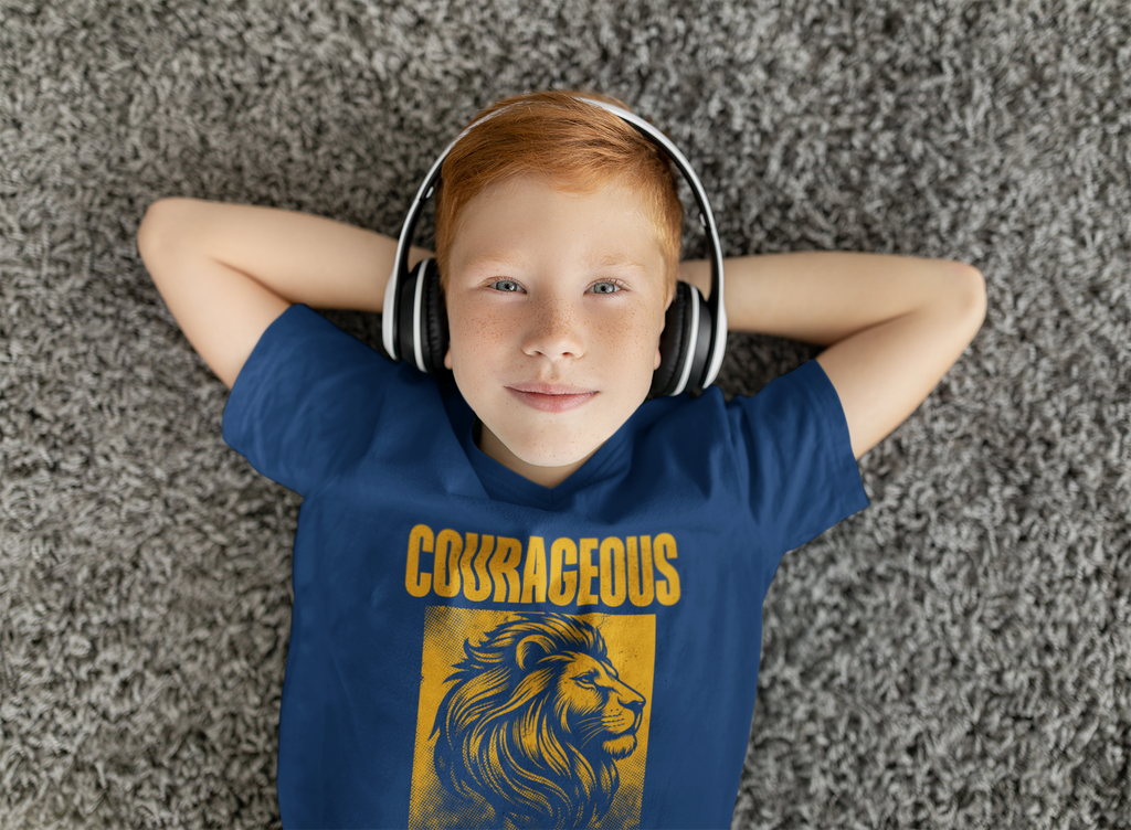 A young boy with red hair lies on a gray textured carpet, wearing a navy blue Christian  t-shirt with a yellow 'COURAGEOUS' lion graphic and white over-ear headphones.