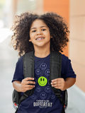 A smiling young kid with curly hair wearing a navy blue faith based graphic t-shirt by SeedSowerGraphics that has a series of smiley faces with a prominent green one in the center with the words below it "Called to be Different" "romans 12:2  and a black backpack, standing outdoors against a blurred orange and beige background