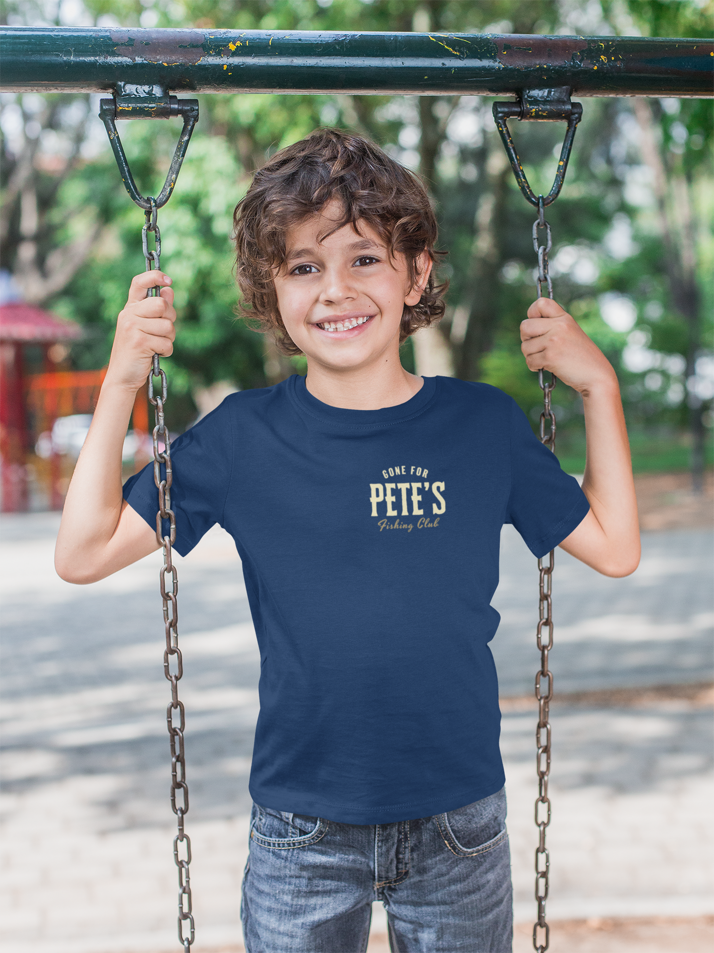 A smiling young boy with curly brown hair stands on a playground swing holding the chains, wearing a navy blue Christian graphic t-shirt with a cream graphic that reads 'GONE FOR PETE'S FISHING CLUB and blue jeans.