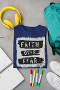 A navy blue Christian graphic t-shirt with a white and black distressed print reading 'FAITH OVER FEAR' and 'Psalm 27:1' is styled in a flat lay on a gray concrete background, surrounded by yellow headphones, a teal backpack, white sneakers, colored markers, and an open notebook.