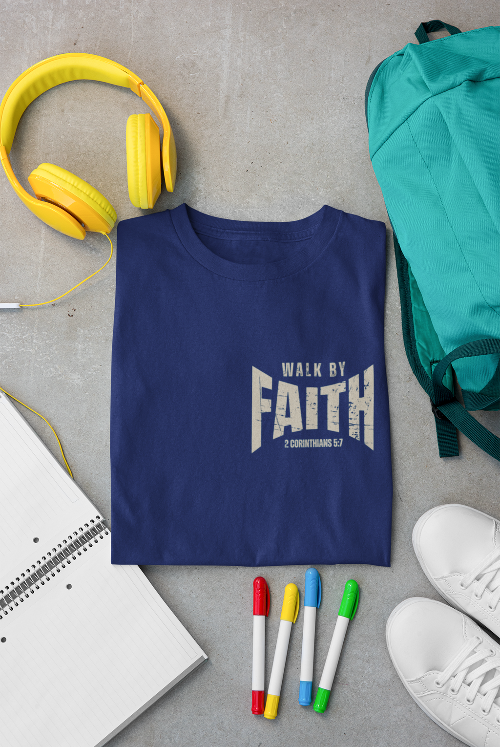 A navy blue Christian graphic t-shirt with a white graphic reading 'WALK BY FAITH' is laid flat on a gray concrete surface, surrounded by lifestyle props including yellow headphones, a teal backpack, a notebook, colored markers, and white sneakers.
