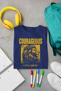 A navy blue graphic Christian youth T-shirt is laid flat on a gray surface, surrounded by school and study props including yellow headphones, a teal backpack, a notebook, four colored markers, and white sneakers. The shirt features a large yellow lion graphic and the words 'COURAGEOUS' and 'BE STRONG. Be COURAGEOUS'.