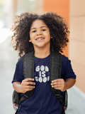 A smiling young kid with voluminous curly hair wears a navy blue Christian graphic t-shirt by SeedSowerGraphics with 'GOD IS GOOD' printed in white, carrying a black backpack. She is photographed from the chest up against a softly blurred background of a warm-toned building.