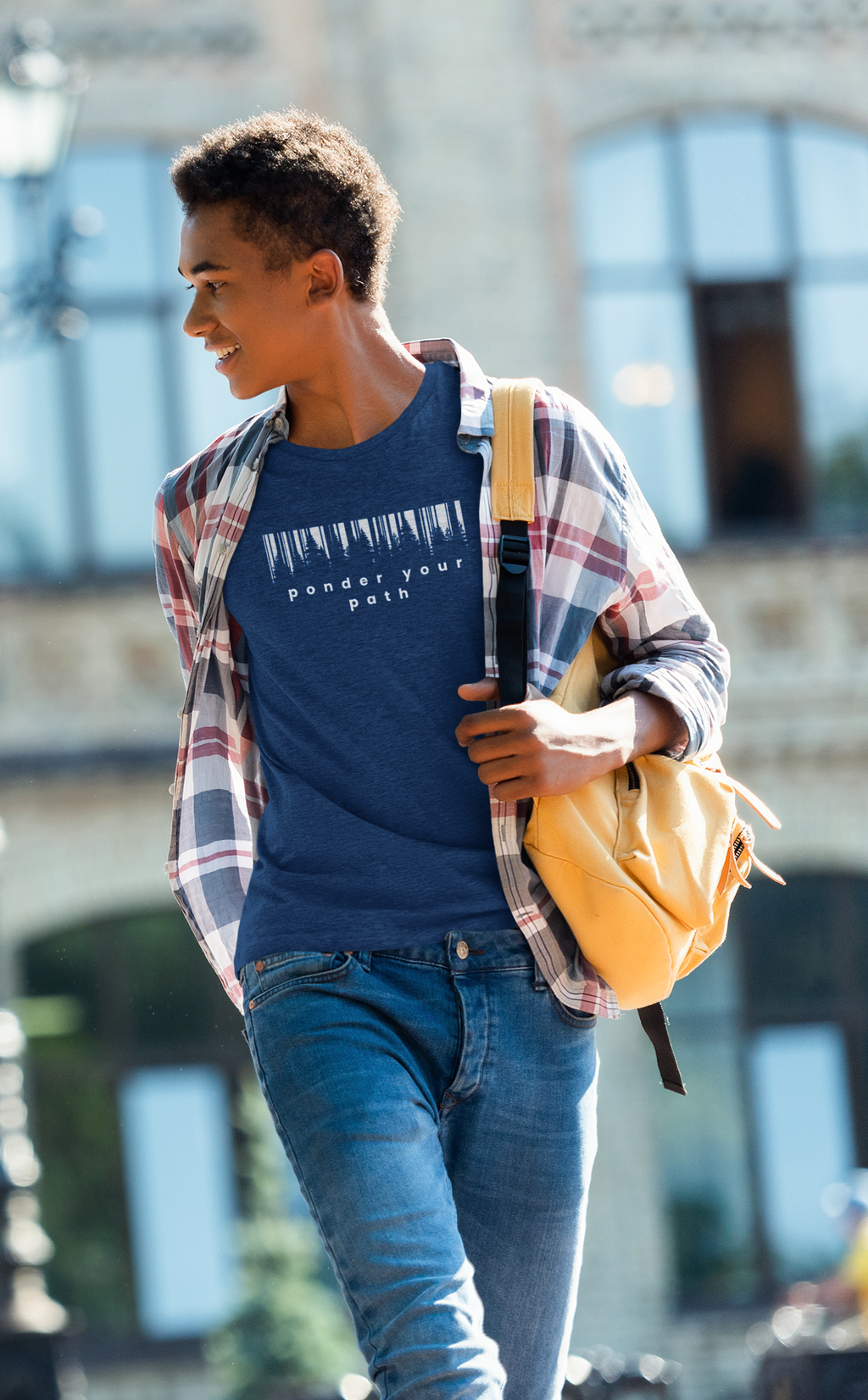 A young man walking outdoors wearing a navy blue Christian graphic t-shirt that says 'Ponder Your Path' under an open red and blue plaid flannel shirt, paired with blue jeans and carrying a yellow backpack.