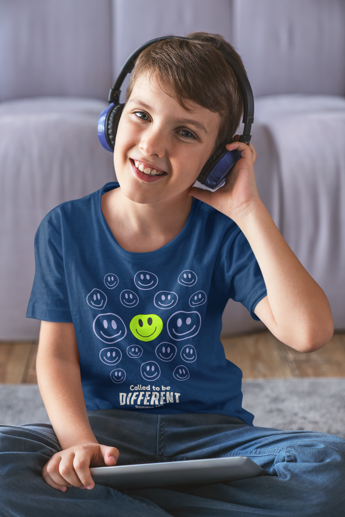 A smiling young boy wearing blue over-ear headphones and a blue graphic faith based T-shirt by SeedSowerGraphics sits cross-legged on a rug in a living room, holding a tablet. The shirt features a green smiley face surrounded by white smiley faces and the text 'Called to be DIFFERENT'.