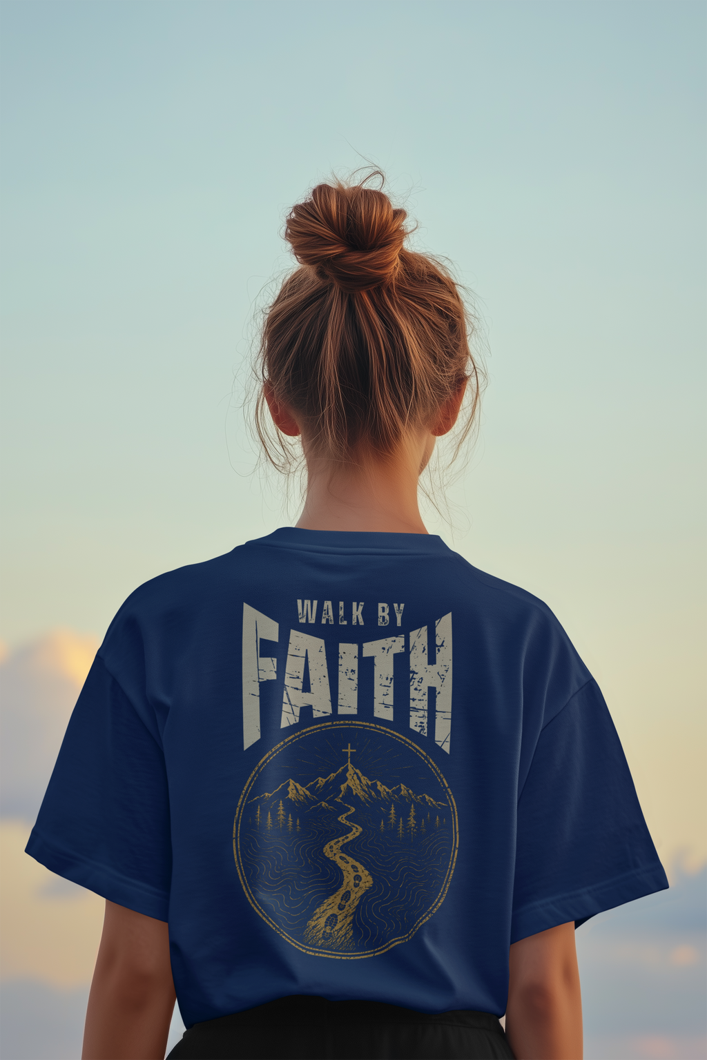 A person viewed from behind wearing a navy blue Christian bible verse T-shirt with a large distressed graphic on the back reading “WALK BY FAITH” above a circular mountain and river illustration, set against a soft sky background.