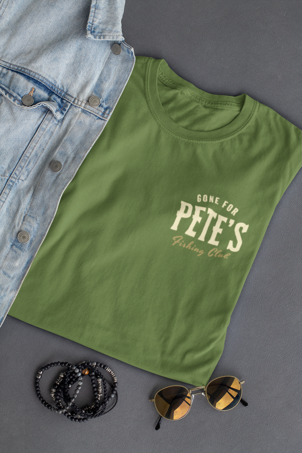 A military green Christian graphic t-shirt with a cream graphic reading 'GONE FOR PETE'S Fishing Club' is styled in a flat lay with a light blue denim shirt, black beaded bracelets, and sunglasses on a dark gray background.