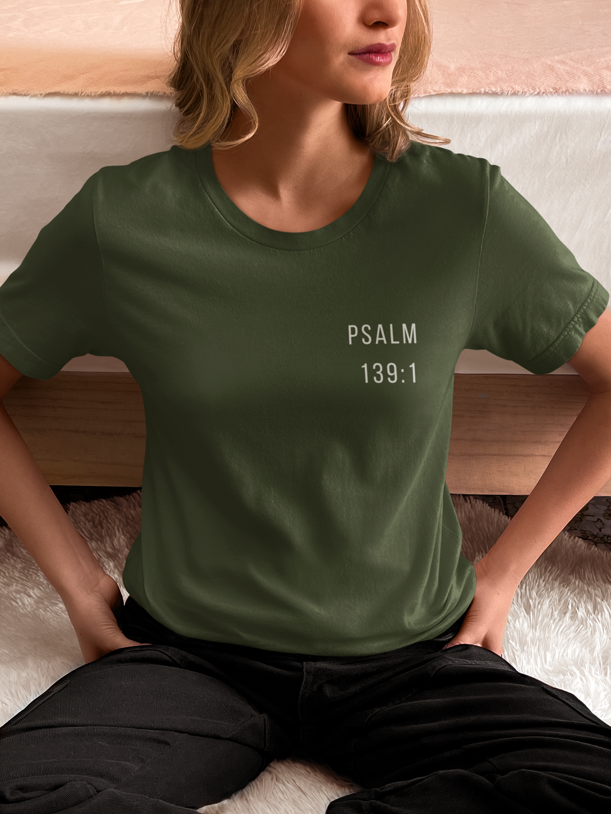 A woman wearing an olive green Christian graphic t-shirt with 'PSALM 139:1' printed in white text on the chest, sitting on a bed.