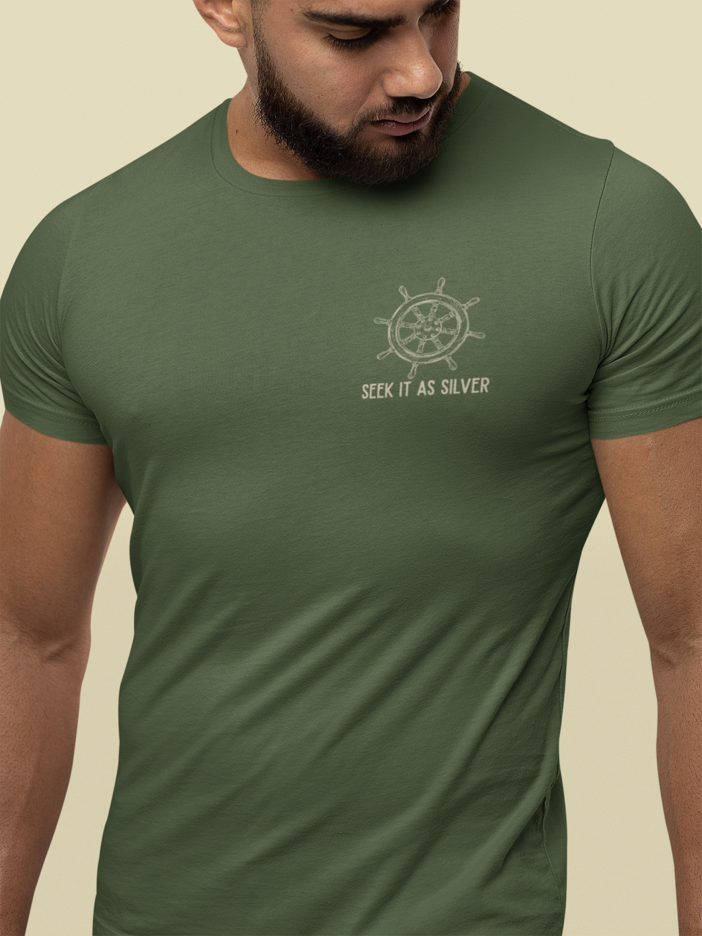 A man wearing an olive green crew-neck Bible verse t-shirt with a white ship's wheel graphic and the text 'SEEK IT AS SILVER' on the chest, set against a solid beige background.