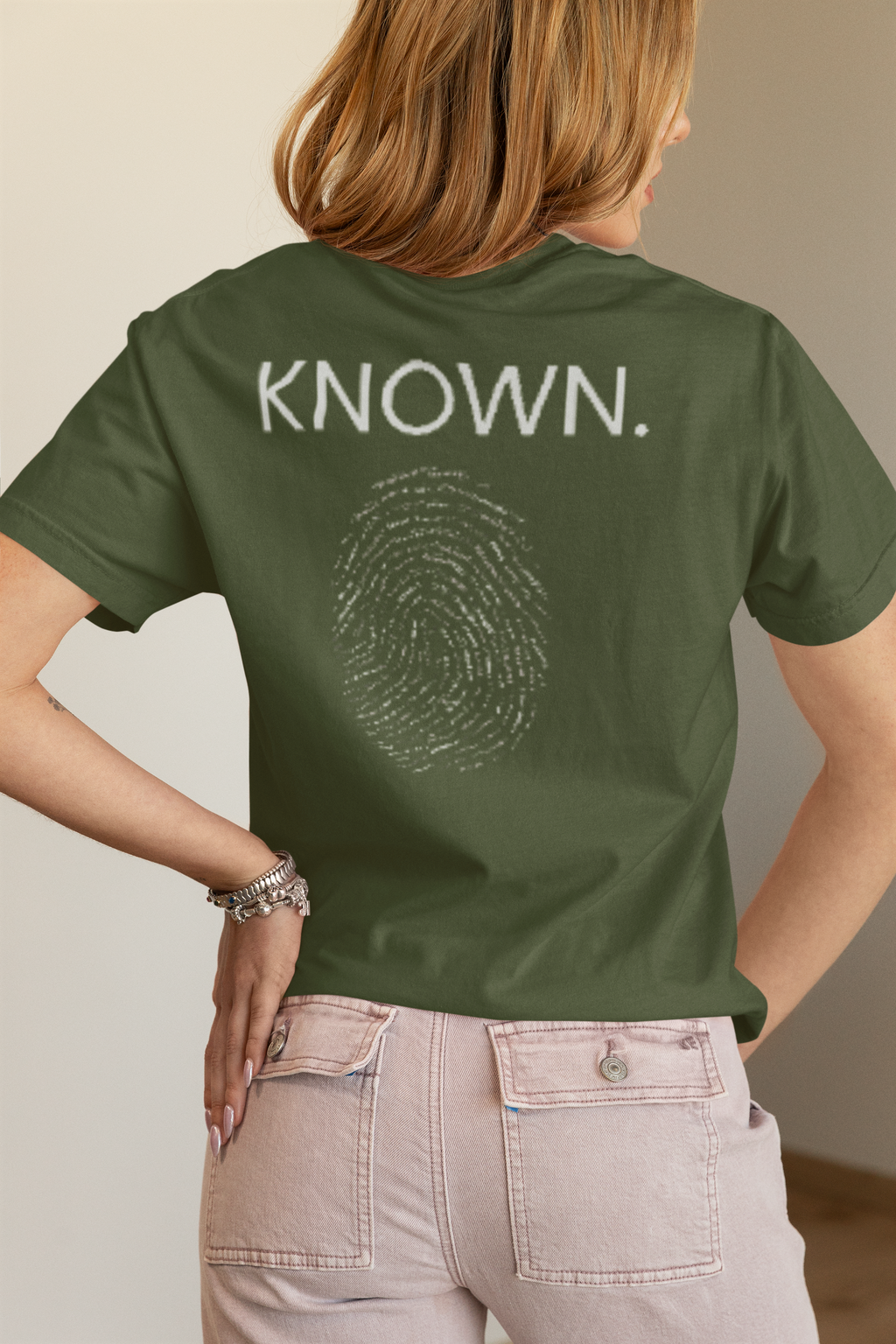 A woman shown from the back wearing an olive-green Christian identity t-shirt with 'KNOWN.' and a fingerprint graphic printed in white on the back, paired with light pink jeans.