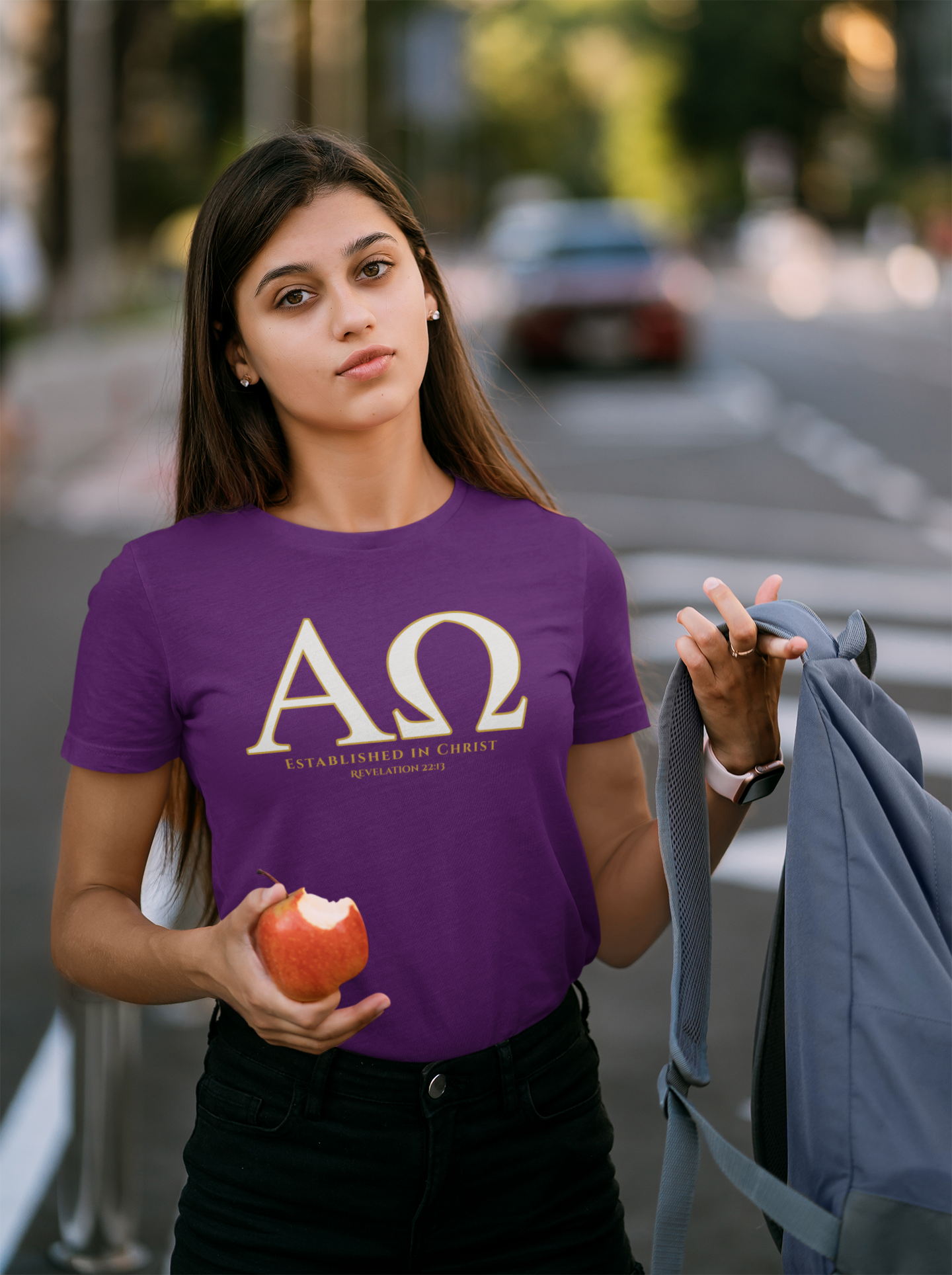A female student wearing a purple faith based t-shirt with a large gold Greek letter 'ΑΩ' (Alpha Omega) and the words 'ESTABLISHED IN CHRIST' 'REVELATION 22:13' printed on the front. She is holding a red apple and a gray backpack, standing outdoors on a blurred street background.