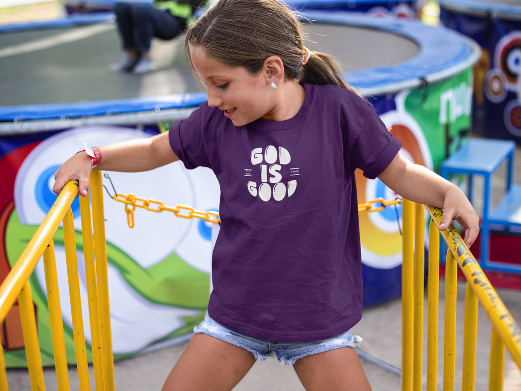 A young girl wearing a purple Christian graphic t-shirt by SeedSowerGraphics with a white graphic that reads 'GOD IS GOOD' stands at an outdoor playground or fair, holding onto yellow safety rails.