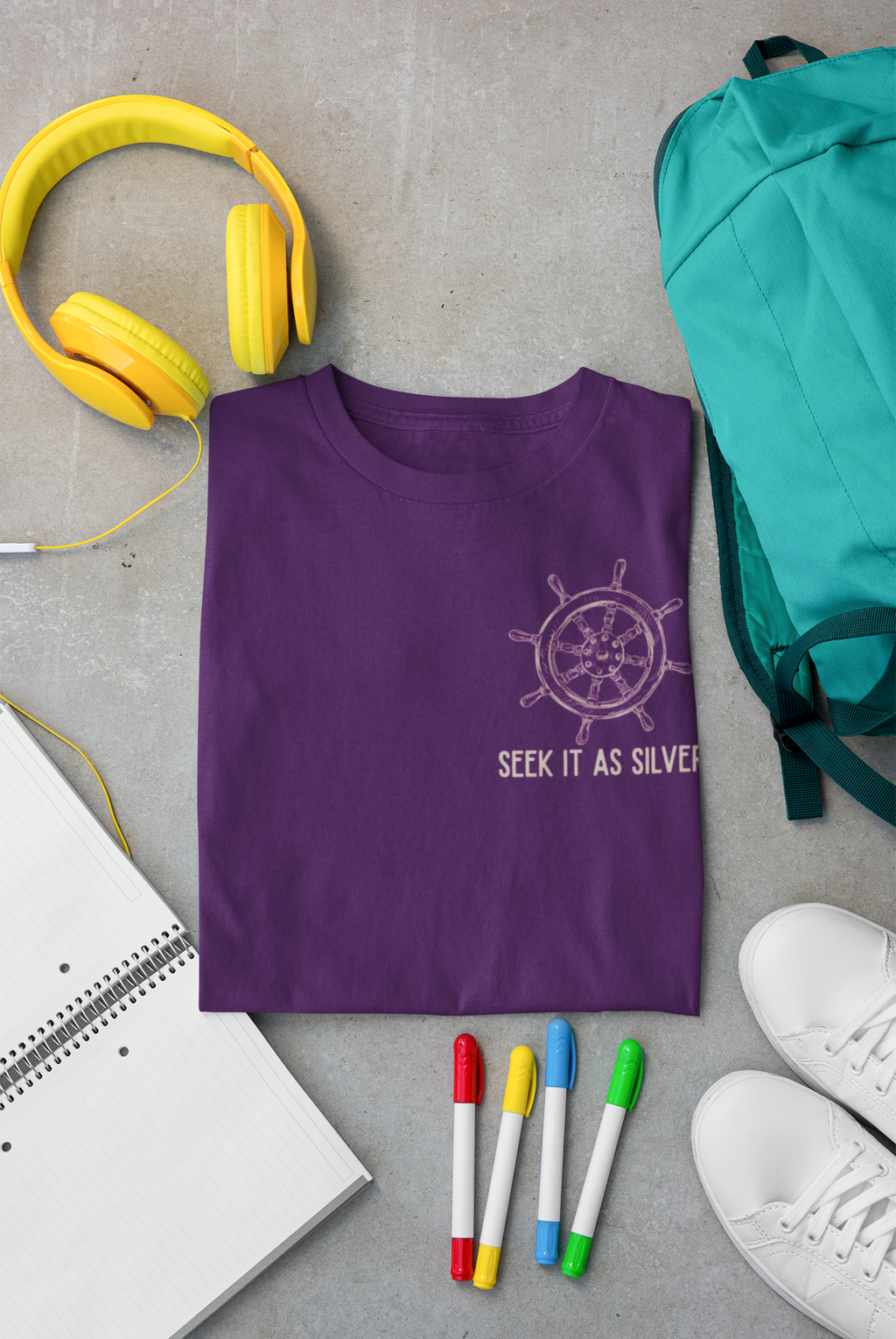 A folded purple Bible verse t-shirt with a silver ship's wheel graphic and the text 'SEEK IT AS SILVER' is displayed in a flat-lay arrangement on a gray concrete background, surrounded by yellow headphones, a teal backpack, a notebook, colored markers, and white sneakers.