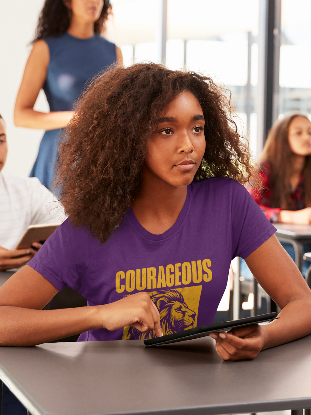 A woman with curly brown hair sits at a desk in a classroom or office setting, wearing a purple Christian graphic T-shirt with the word 'COURAGEOUS' and a lion illustration in yellow. She is holding a tablet, with other people blurred in the background.