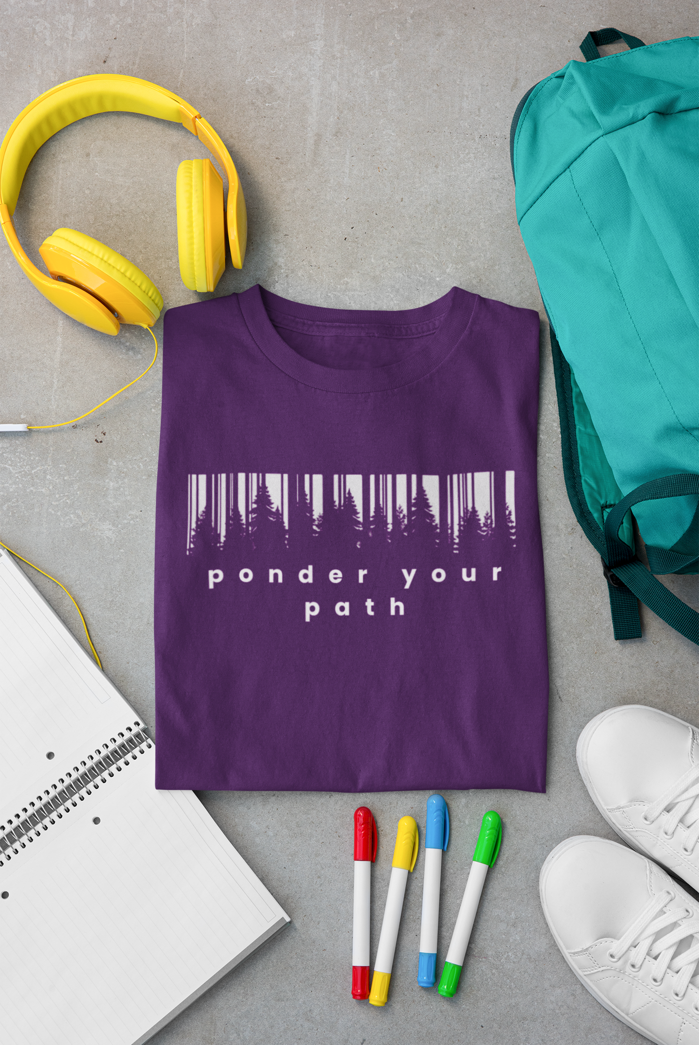 A folded purple  youth Chrisitan T-shirt with a white forest silhouette and the words 'ponder your path' printed on the front, styled in a flat lay with yellow headphones, a teal backpack, a notebook, four colored markers, and white sneakers on a gray concrete surface.