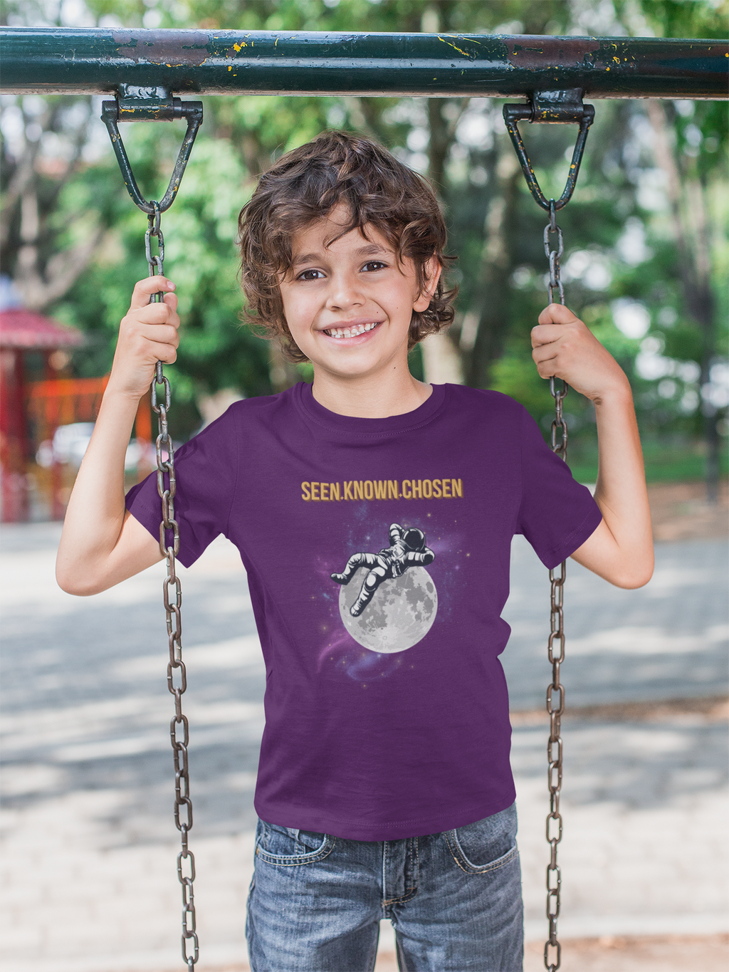 A smiling young boy stands on a playground swing holding the chains, wearing a purple Christian graphic t-shirt with the text 'SEEN.KNOWN.CHOSEN'