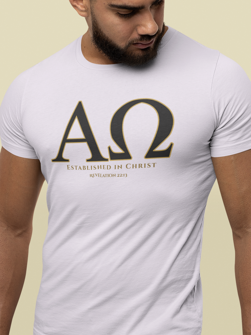 A man wearing a white Bible verse t-shirt with a large black Greek letter 'ΑΩ' (Alpha Omega) graphic outlined in gold, with the text 'ESTABLISHED IN CHRIST' and 'REVELATION 22:13' printed below.