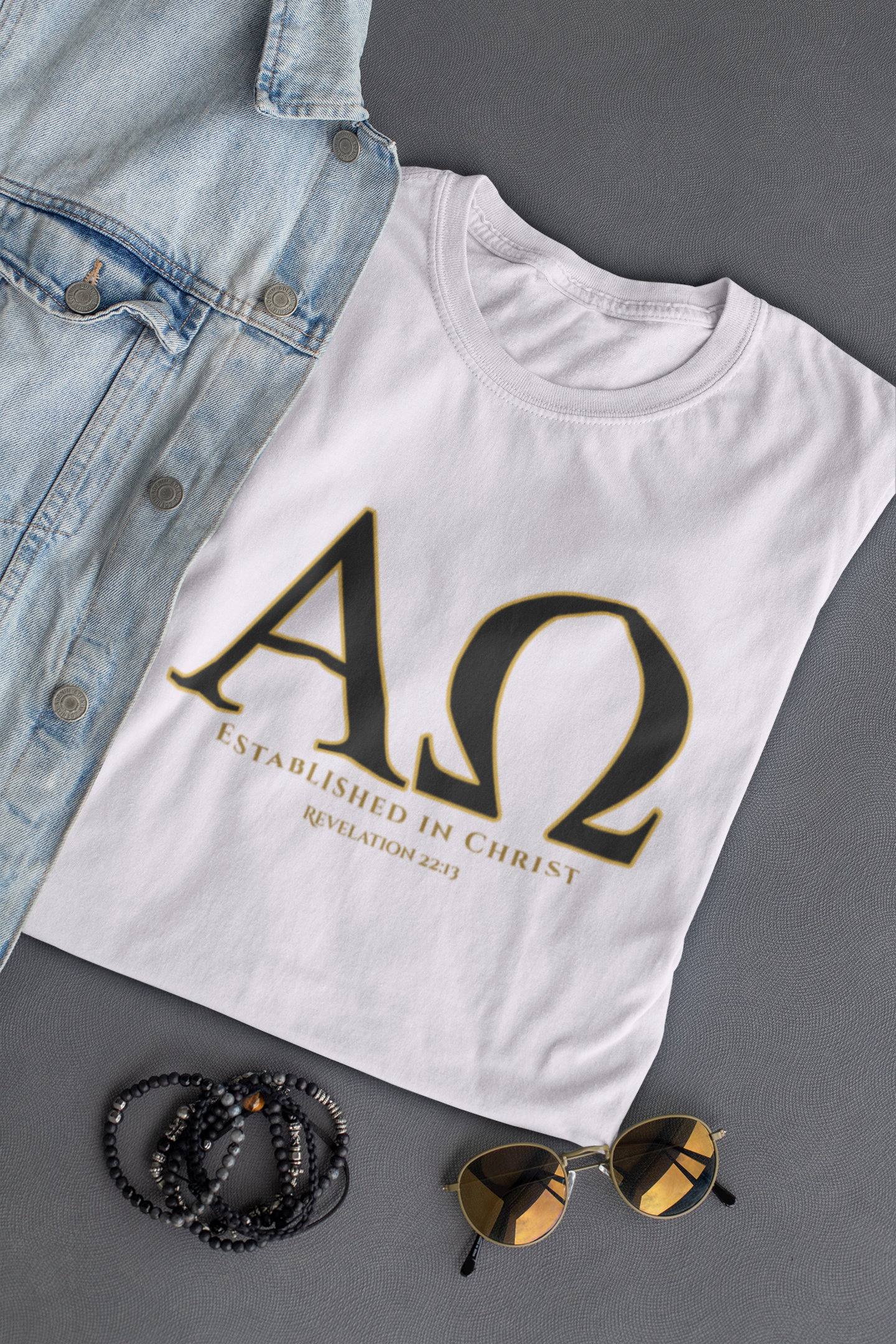 A white Christian based T-shirt laid flat on a dark gray surface, styled with a light blue denim jacket, black beaded bracelets, and gold-tinted sunglasses. The shirt features a large black Greek letter design Alpha Omega with gold outlining and the text 'ESTABLISHED IN CHRIST' and 'REVELATION 22:13'.