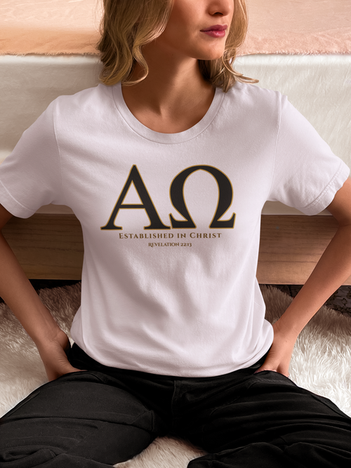 A woman wearing a white Christian Scripture T-shirt with a large black Greek letter 'ΑΩ' and the text 'ESTABLISHED IN CHRIST' and 'REVELATION 22:13' printed on the front, paired with black pants, sitting on a white rug in front of a bed.