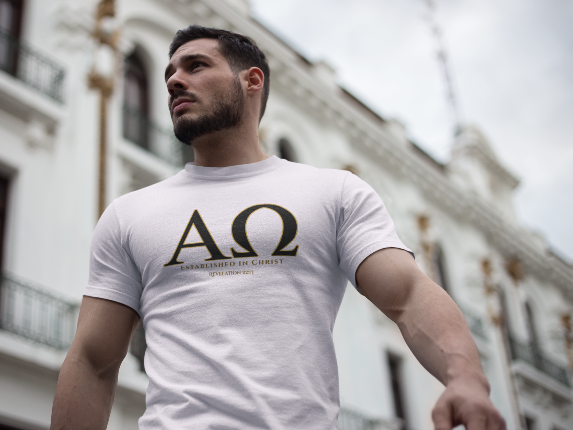 A man wearing a white Christian graphic t-shirt with a large black Greek letter 'ΑΩ' (Alpha Omega) and the words 'ESTABLISHED IN CHRIST' 'REVELATION 22:13' printed on the chest, photographed outdoors against a blurred white building background.