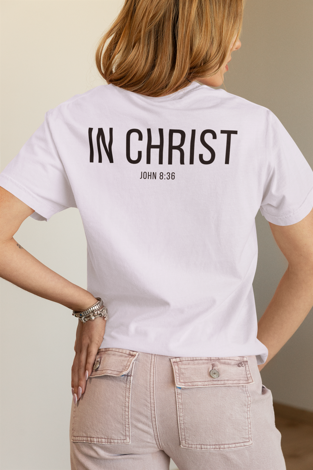 A woman shown from the back wearing a white minimalist Christian T-shirt with the text 'IN CHRIST' and 'JOHN 8:36' printed in black on the back, paired with light beige jeans.
