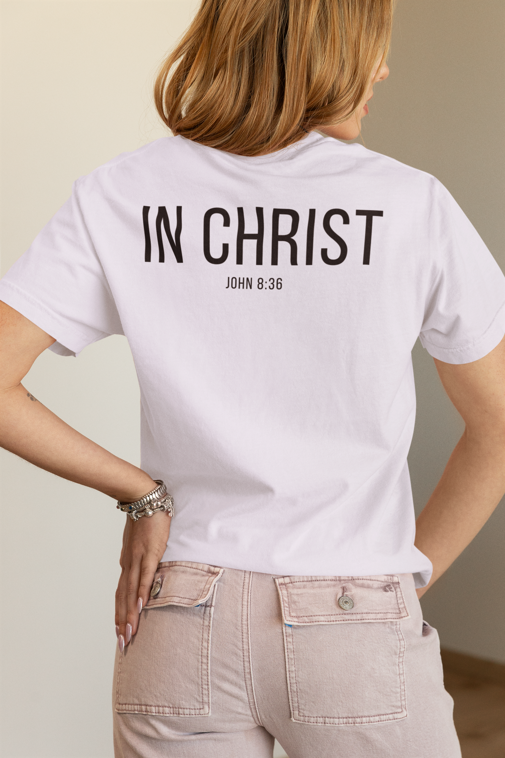 A woman shown from the back wearing a white minimalist Christian T-shirt with the text 'IN CHRIST' and 'JOHN 8:36' printed in black on the back, paired with light beige jeans.