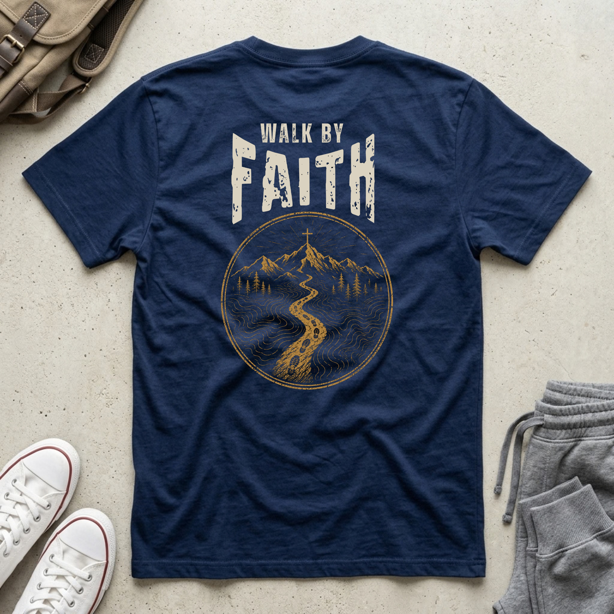 A navy blue t-shirt laid flat on a light concrete surface, showing the back with a distressed white 'WALK BY FAITH' text and a gold circular mountain and river graphic. White sneakers, a brown backpack, and gray sweatpants are partially visible around it as styling props.