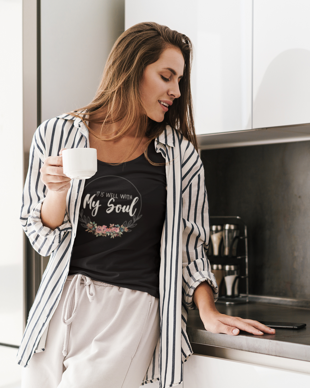 A woman in a kitchen setting wearing a black Christian graphic t-shirt by SeedSowerGraphics with 'It is well with My Soul' written on it, layered under an open black and white striped button-up shirt and paired with light beige drawstring pants. She is holding a white mug.