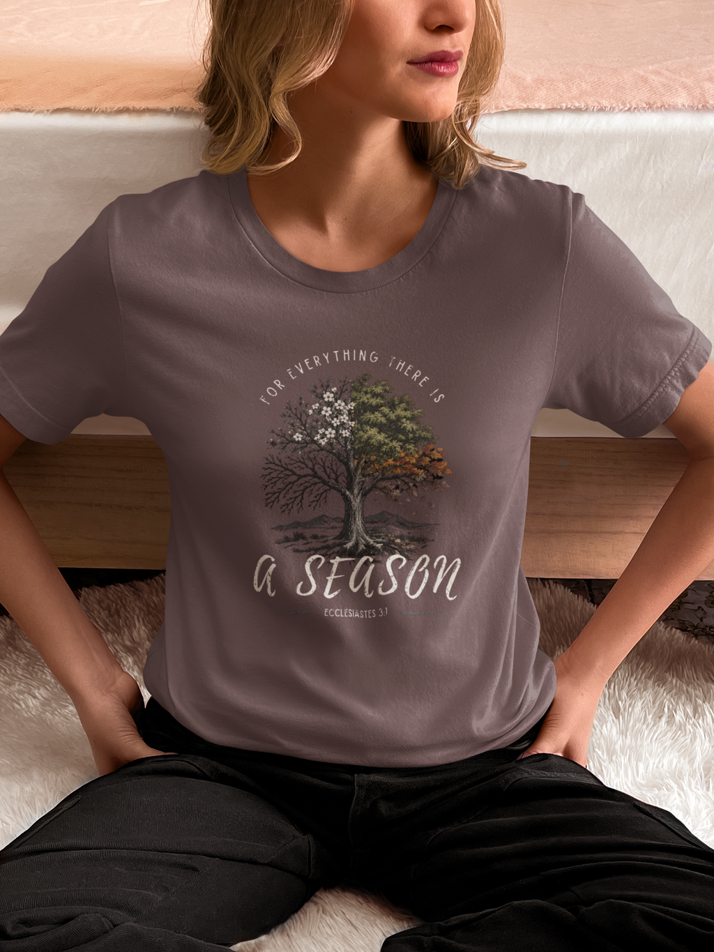 A woman wearing a taupe-gray Christian graphic t-shirt by SeedSowerGraphics with a tree design and the text 'FOR EVERYTHING THERE IS A SEASON' and 'Ecclesiastes 3:1', sitting on a white rug in front of a bed.