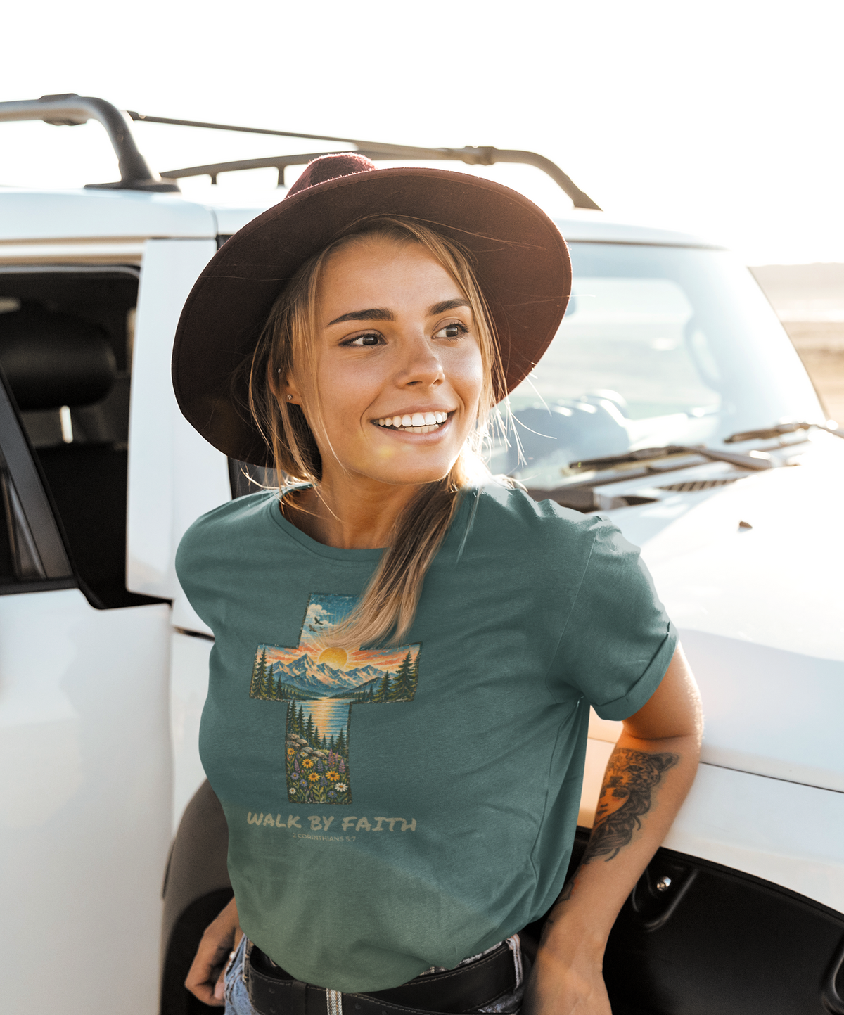 A woman wearing a dark brown wide-brim hat and a sage green Christian graphic t-shirt with a cross and mountain design, posing next to a white vehicle outdoors.