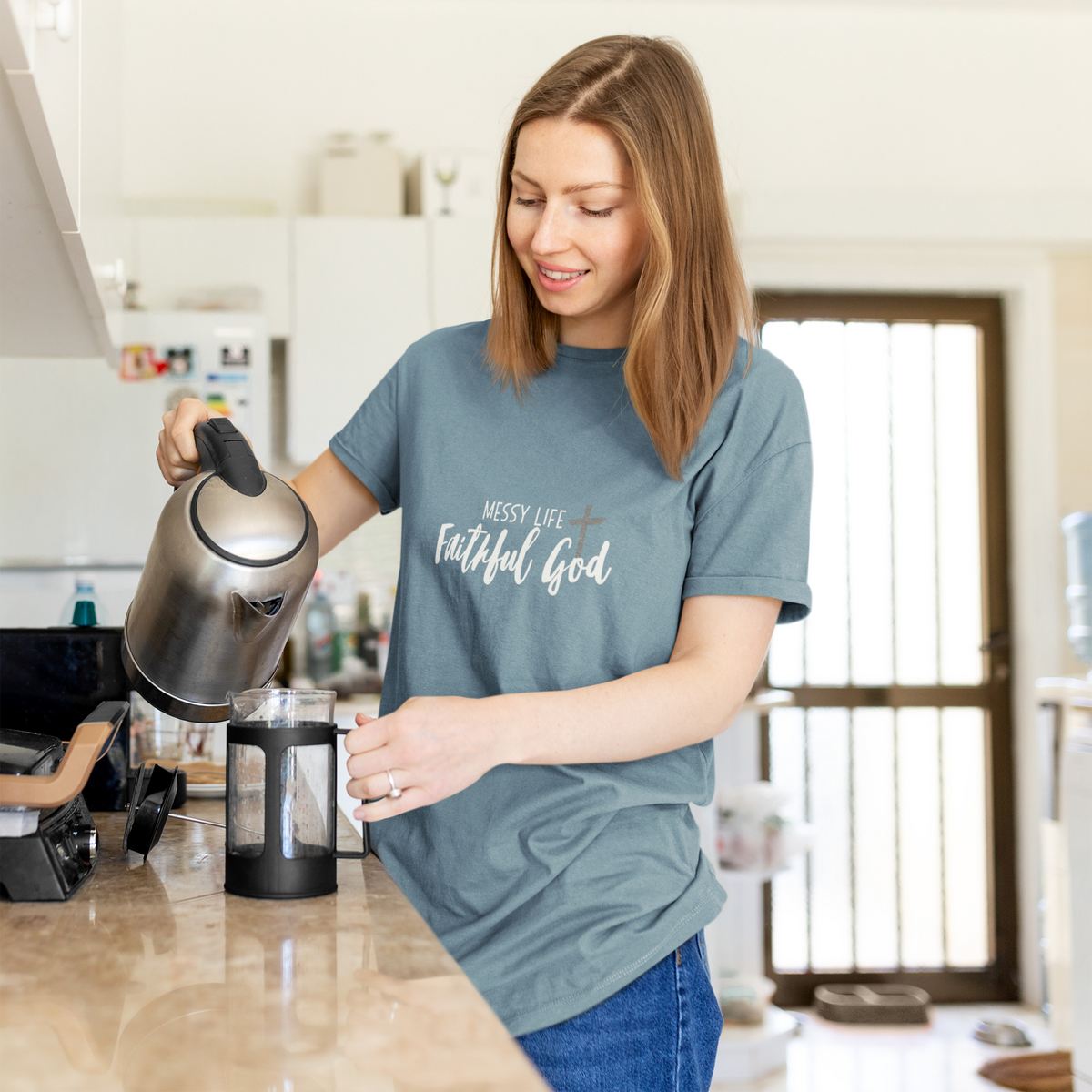 A woman in a kitchen wearing a slate blue faith based t-shirt by SeedSowerGraphics with the text 'MESSY LIFE Faithful God' in white script. She is pouring water from a stainless steel kettle into a French press.