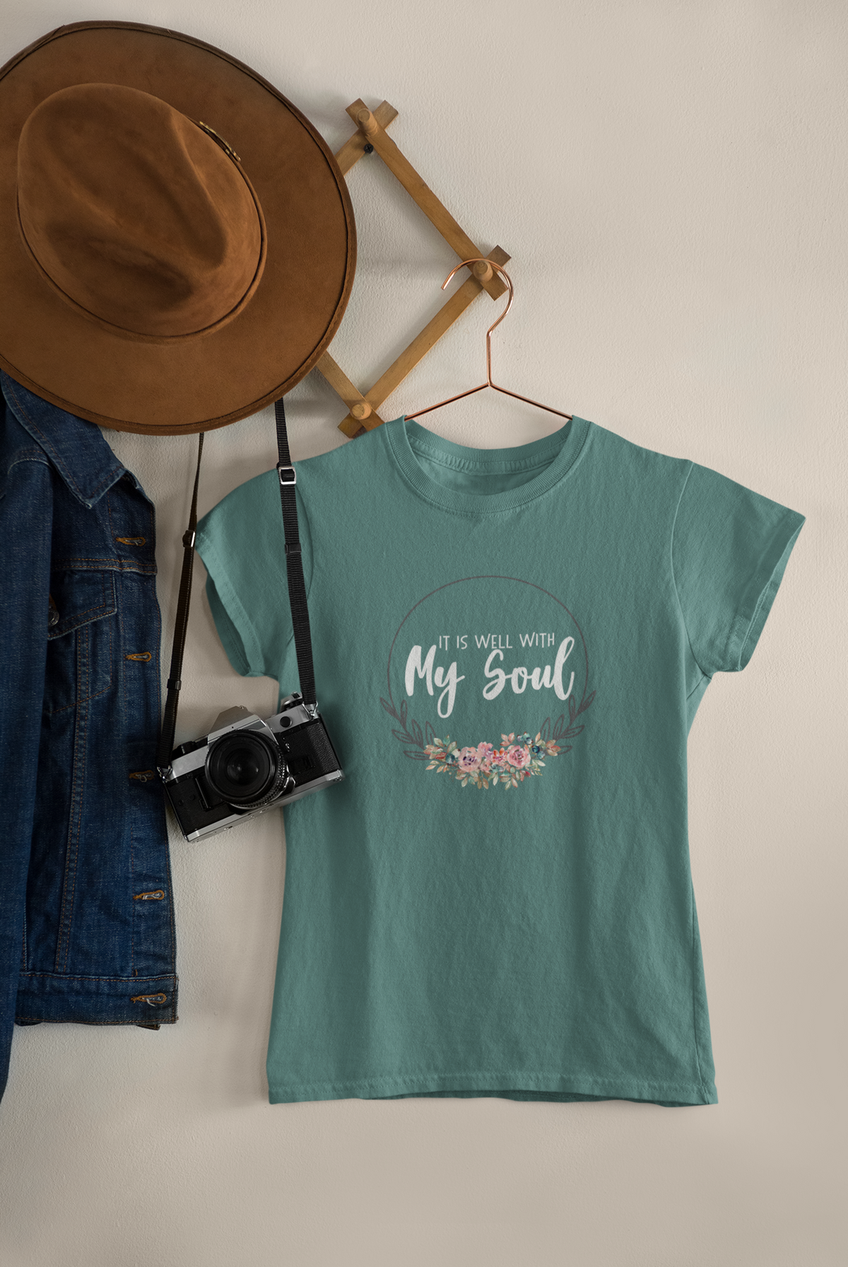 A teal short-sleeve Christian graphic T-shirt by SeedSowerGraphics hanging on a wooden hanger against a light beige wall, styled with a brown wide-brim hat, a denim jacket, a vintage camera, and a wooden wall rack.