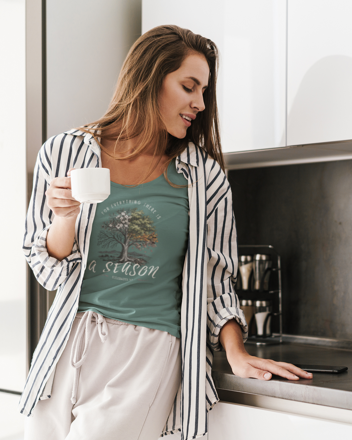 A woman in a kitchen wearing a sage green Christian graphic top by SeedSowerGraphics  that reads 'For everything there is a season' under an open black-and-white striped shirt. She is holding a white mug and leaning on a stainless steel counter.