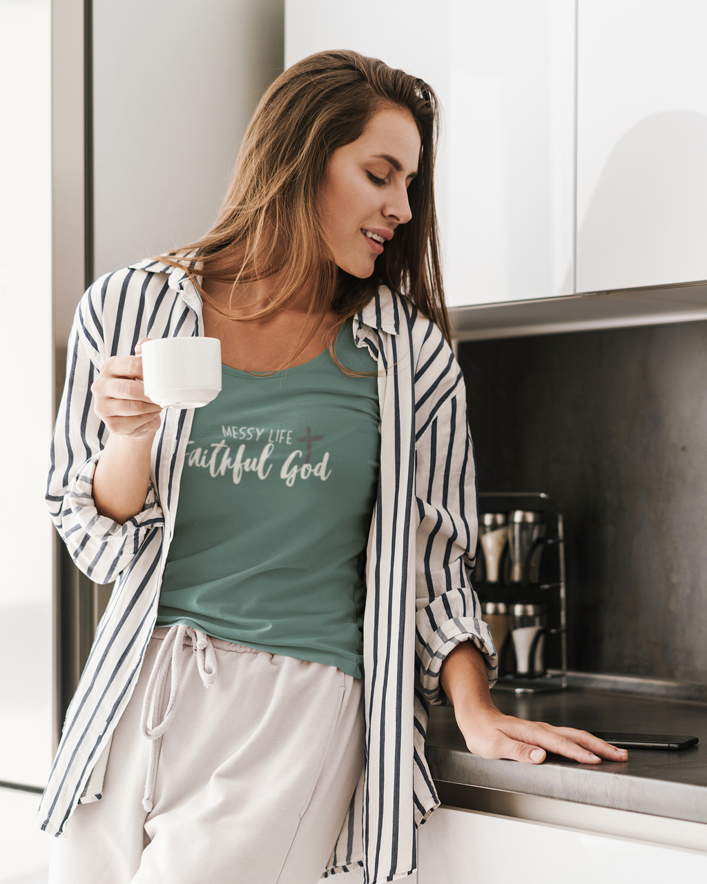 A woman in a kitchen wearing a sage green Christian graphic shirt by SeedSowerGraphics  that reads 'Messy Life + Faithful God' under an open black-and-white striped shirt. She is holding a white mug and leaning on a stainless steel counter.